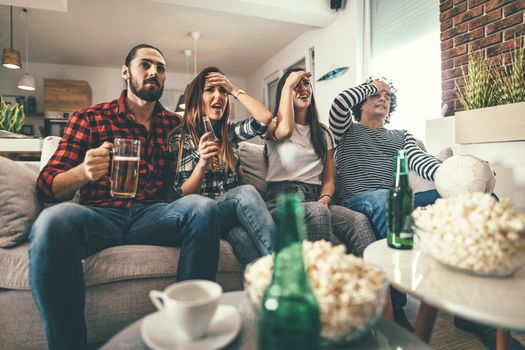 Friends are fans of sports games as football love spending their free time at home together. They are screaming and gesturing for a victory. 