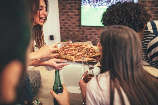 Happy friends having fun looking TV at home sitting on sofa, while eating pizza and drinking beer. They have great weekend in nice company indoors. 