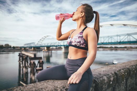 Young fitness woman is resting after hard training on the wall by the river bridge and drinking water.