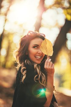 Cute young woman enjoying in sunny forest in autumn colors. She is holding golden leaf and looking at camer.