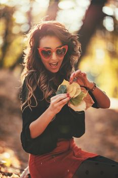 Cute young woman enjoying in sunny forest in autumn colors. She is holding golden yellow leaves.