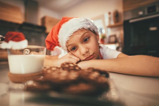 A little pensive girl looks in a cup of milk and plate with a cookies and waiting Santa Clause.