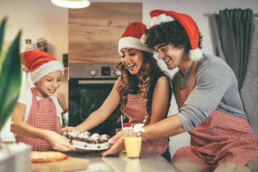 Happy parents and their daughter with santa's cap are preparing meal together in the kitchen. Little girl is holding plate with muffin offering to her mother and father.