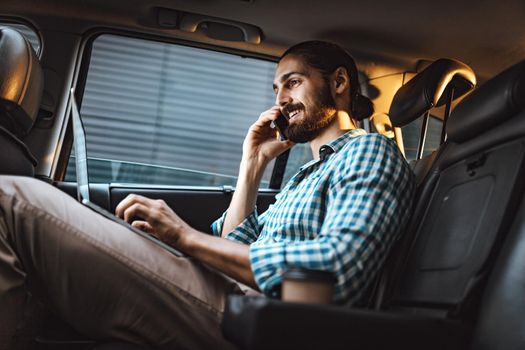 Young confident businessman is holding smartphone in his hand and texting on a laptop on the back seat in car.