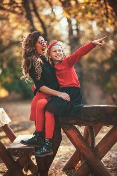 Beautiful young mother and her daughter walking through forest. Little girl is sitting on the wooden picnic table and pointing with finger far away.