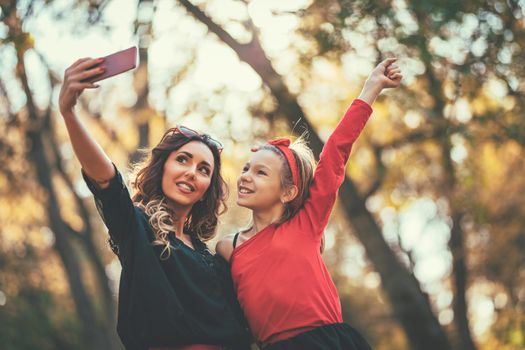 Beautiful young mother and her happy daughter having fun in the forest in sunset. They are  smiling and having fun with smartphone taking selfie.