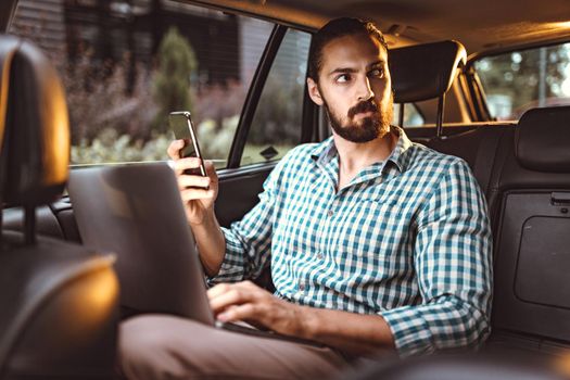 Young confident businessman is holding smartphone, talking and working on a laptop on the back seat in car.