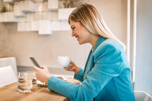 Young smiling businesswoman on a break in a cafe. He is drinking coffee and using smartphone. 