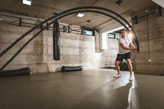 Young man doing strength training using heavy ropes at the gym. Athlete moving the ropes in wave motion as part of strength training.