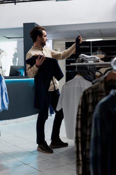 Clothing store customer selecting tie and shirt while shopping for formal outfit. Young man exploring fashion boutique while holding garment and accessory, checking style