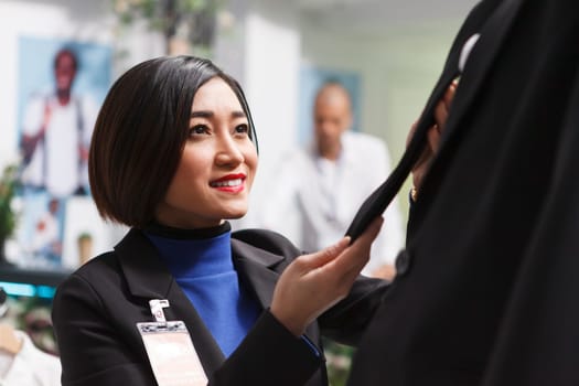 Smiling asian woman seller checking formal jacket fit on mannequin while organizing merchandise in clothing store. Fashion boutique employee adjusting trendy suit for sale