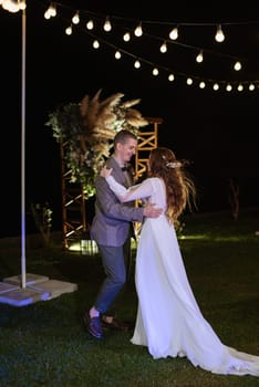 the first wedding dance of the bride and groom in the glade of the country club in the light of sunset and warm garlands