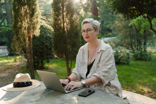 woman freelancer in glasses works at a computer at a white table in nature and spends her day productively.