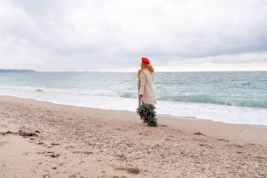 Redhead woman Christmas tree sea. Christmas portrait of a happy redhead woman walking along the beach and holding a Christmas tree in her hands. She is dressed in a light coat and a red beret