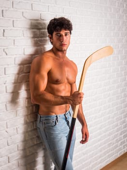 Handsome young man shirtless with hockey stick in hands leaning against white brick wall, looking at camera, wearing jeans