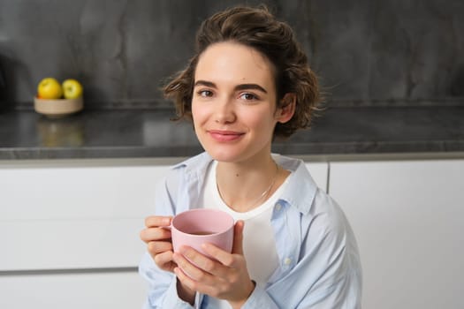 Portrait of smiling, beautiful young woman, drinking coffee in kitchen, morning magic with cuppa tea, looking tenderly.