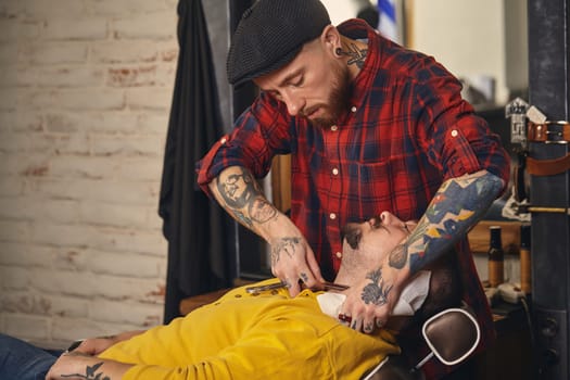 Client with beard and moustache sit on chair, and professional barber make beard shaving in barber shop