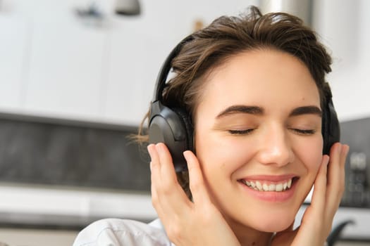 Close up portrait of cute young woman, smiling and listening to music in wireless headphones, enjoying good sound quality.