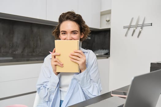 Portrait of smiling brunette girl, holds her planner, writes in notebook and looks happy, laughs, does homework, makes notes, sits at home in kitchen.