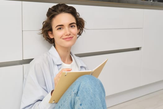 Portrait of creative young woman, artist sits on floor and draws in her planner, writes diary, smiling while doing art at home in cozy space.