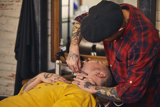 Client with beard and moustache sit on chair, and professional barber make beard shaving in barber shop