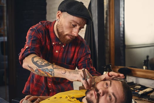 Client with beard and moustache sit on chair, and professional barber make beard shaving in barber shop