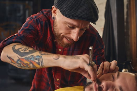 Client with beard and moustache sit on chair, and professional barber make beard shaving in barber shop