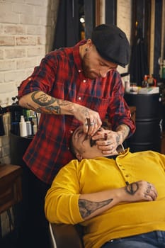 Client with beard and moustache sit on chair, and professional barber make beard shaving in barber shop