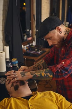 Client with beard and moustache sit on chair, and professional barber make beard shaving in barber shop