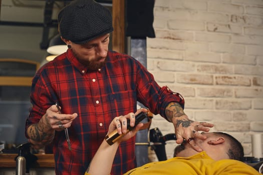 Client with beard and moustache sit on chair, and professional barber make beard shaving in barber shop