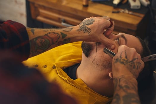 Client with beard and moustache sit on chair, and professional barber make beard shaving in barber shop