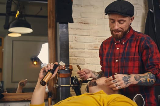 Client with beard and moustache sit on chair, and professional barber make beard shaving in barber shop