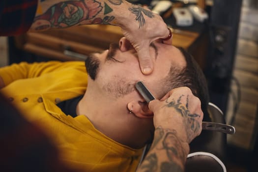 Client with beard and moustache sit on chair, and professional barber make beard shaving in barber shop