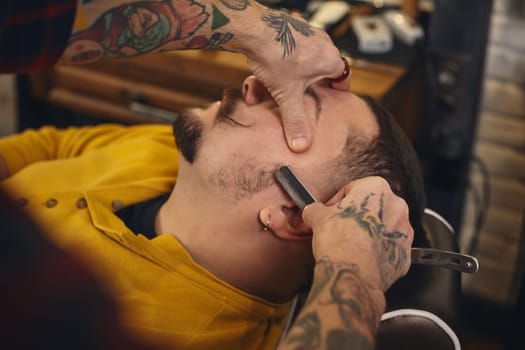 Client with beard and moustache sit on chair, and professional barber make beard shaving in barber shop