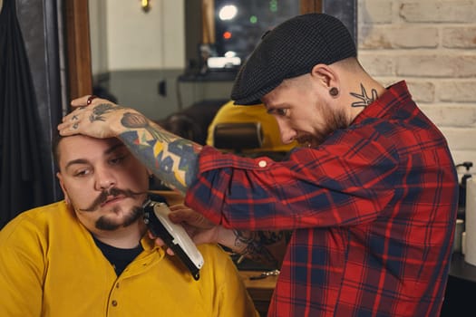 Client with beard and moustache sit on chair, and professional barber make beard shaving in barber shop