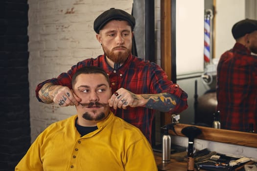 Client with beard and moustache sit on chair, and professional barber make beard shaving in barber shop. The barber straightens the mustache of the client
