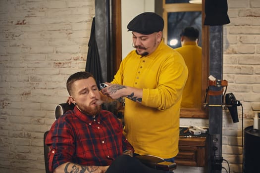 Barber with mustache making haircut of attractive bearded man in barbershop with trimmer