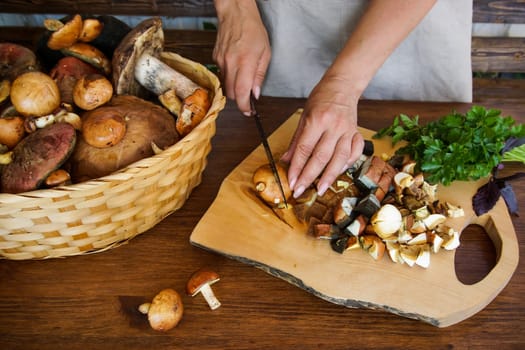 Women's hands cut wild mushrooms on a wooden board. There is a basket of mushrooms on the table and fresh greens.