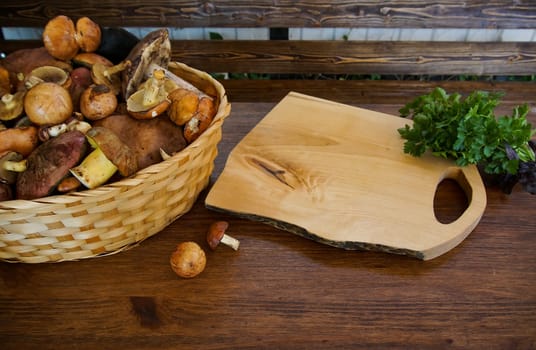 preparation of forest edible mushrooms. There is a basket of mushrooms on the table, there is a cutting board and there are fresh greens