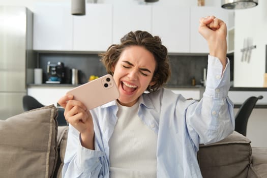Image of girl celebrating, holding smartphone and cheering from excitement, winning, receive great news on mobile phone. People and lifestyle concept