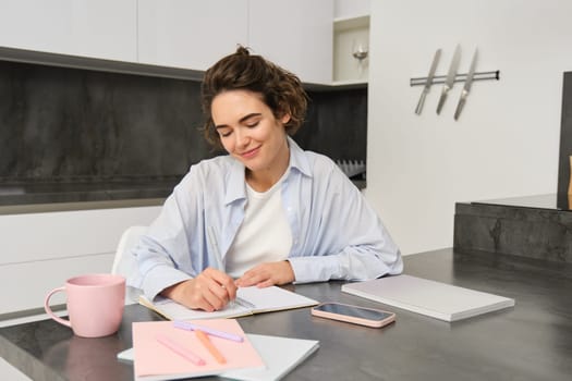 Portrait of young woman working from home, writing down information in notebook, taking notes, sitting in kitchen and studying, student doing homework.