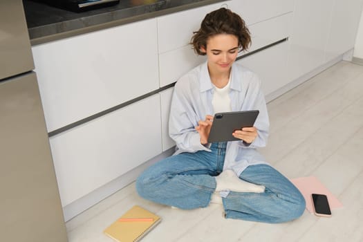 Young woman, freelancer works from home, looks at her digital tablet, reads through documents online, smiling and sitting on floor in kitchen.