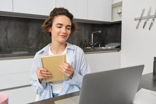 Portrait of businesswoman works from home, looks at laptop and writes down information, makes notes, studies online on course website.