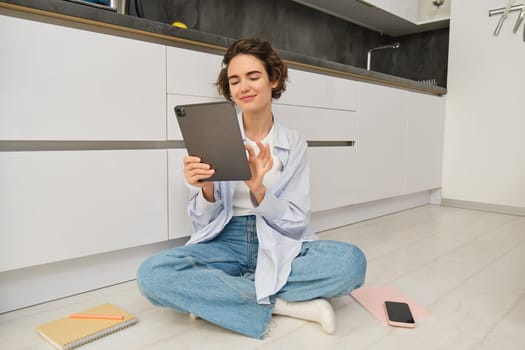 Image of young woman looking at her digital tablet, sits on floor with documents, work papers and smartphone, working from home, studying online, reading ebook.