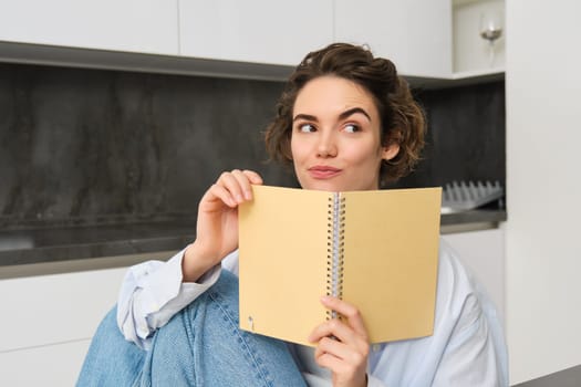 Portrait of beautiful young woman, looking intrigued, reading notes and smiling with thoughtful face, sitting at home in kitchen. Lifestyle and people concept