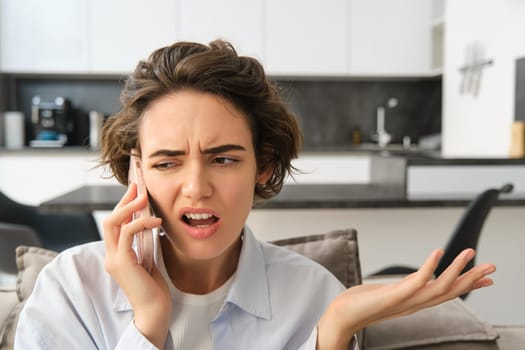 Image of woman with confused, annoyed face, talking on mobile phone while sitting at home on sofa, shrugging and looking bothered.