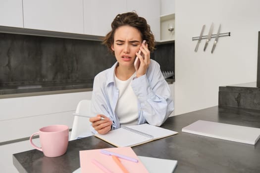 Portrait of woman working on remote, calling someone and taking notes, looks confused, hearing something complicated during mobile conversation.