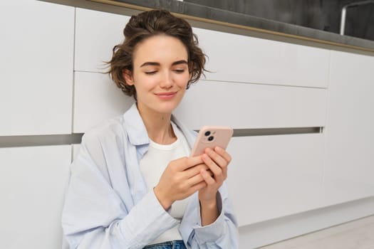 Indoor shot of happy girl with smartphone, sits on floor at home and uses mobile phone, watches video in app, chats with friends in applicatiion.