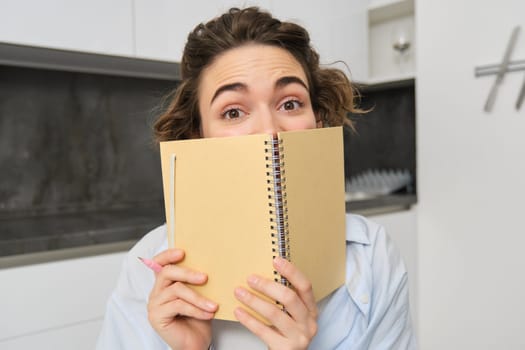 Portrait of happy young woman, hides her face behind planner, shows her diary notebook and smiles at camera, works from home.