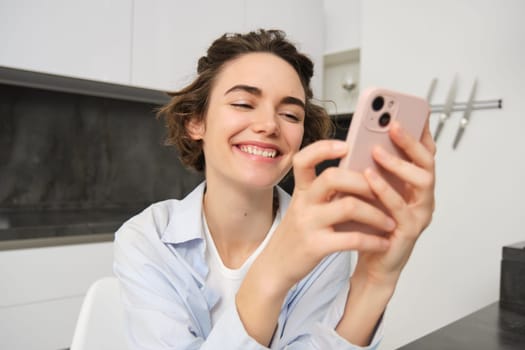 Portrait of brunette woman chats on mobile phone, uses smartphone app from home, orders delivery while sits in kitchen.